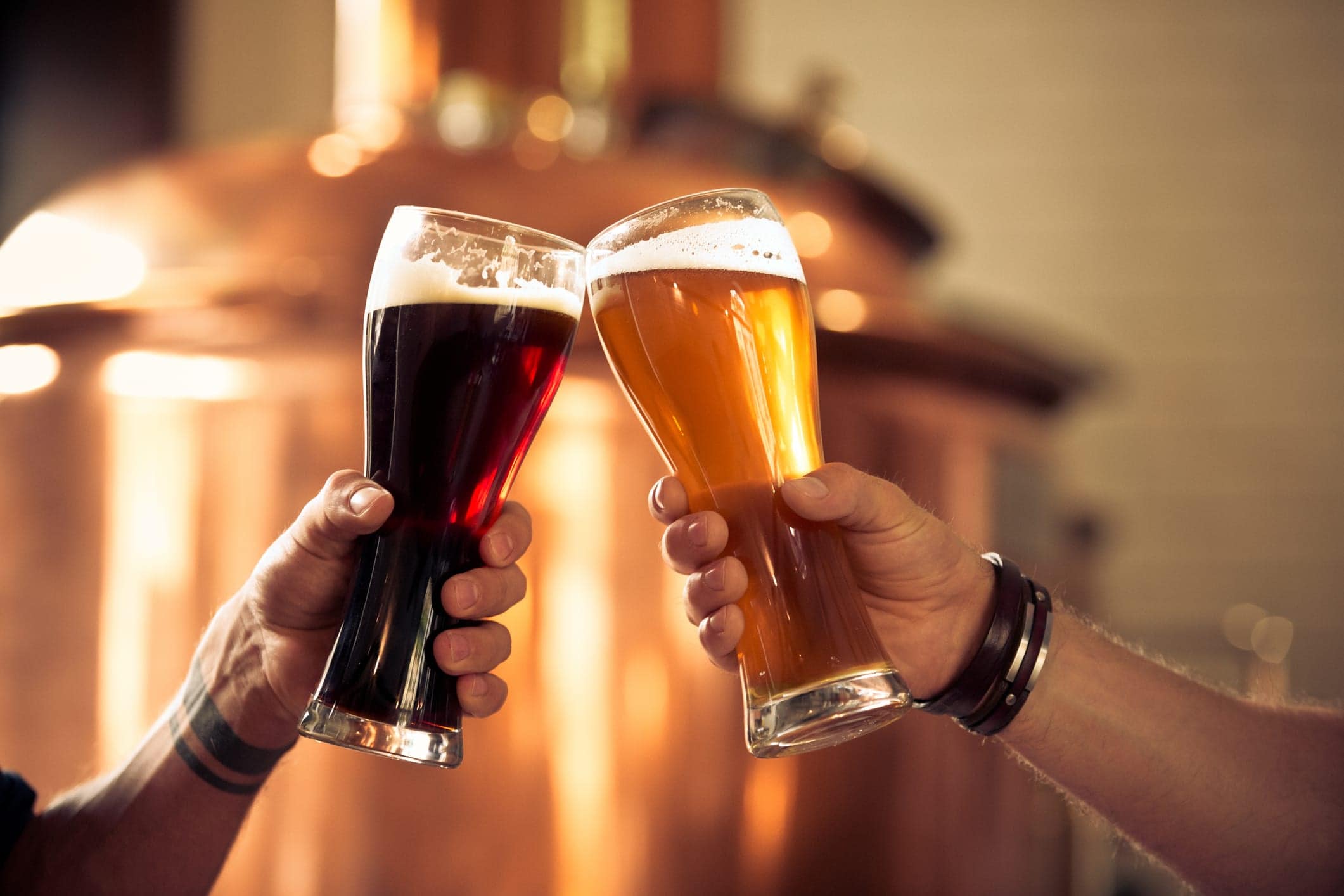 Friends toasting with beer glasses in the microbrewery. Close up of pint glasses and hands, unrecognizable people. Copper vat in the background.