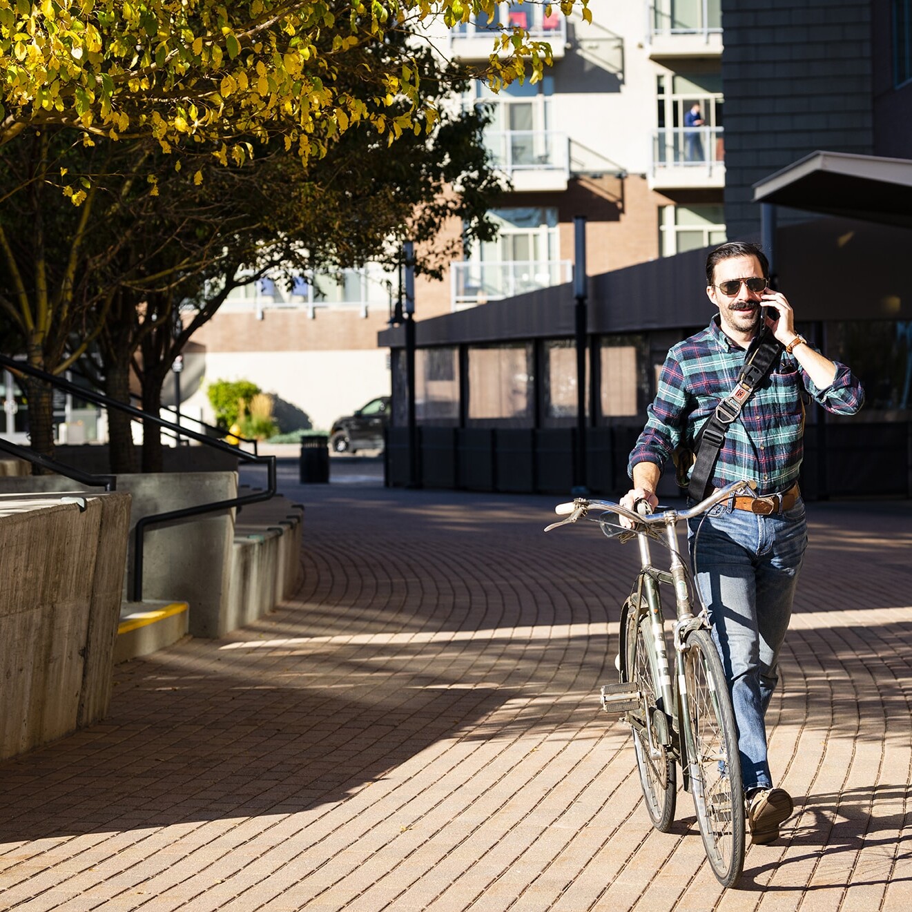 A person walking a bicycle while talking on a phone, set in an urban environment with trees and modern buildings in the background.