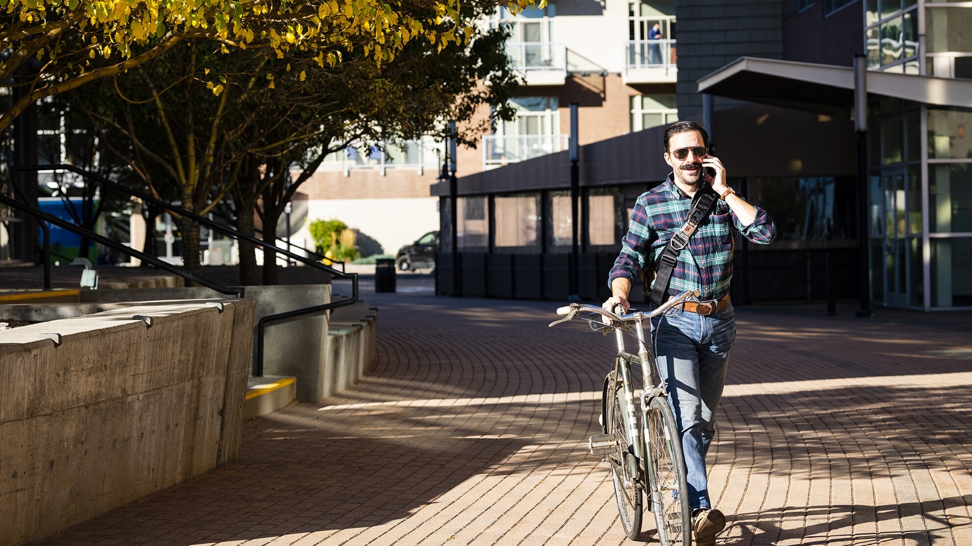 A person walking a bicycle while talking on a phone, set in an urban environment with trees and modern buildings in the background.