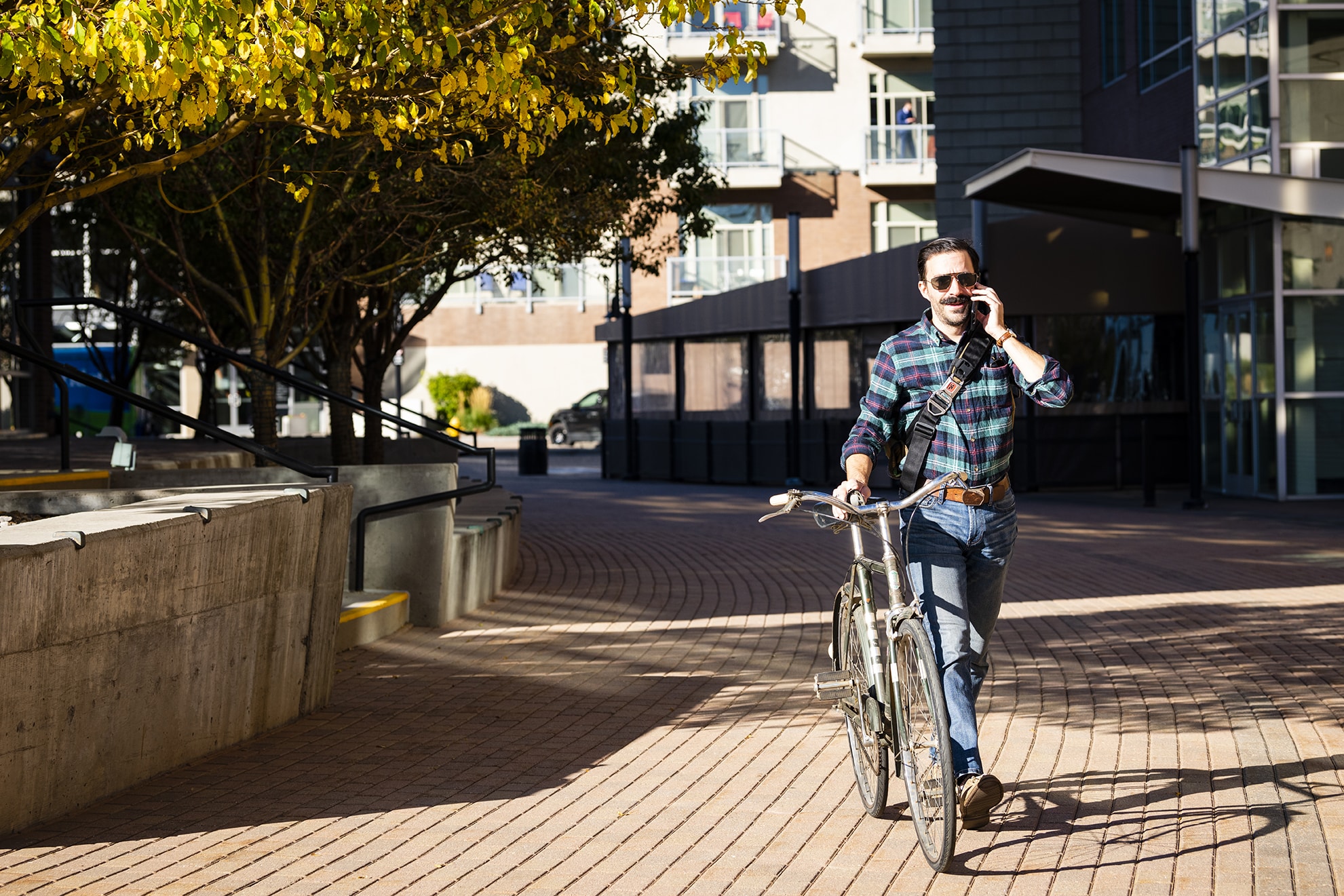 A person walking a bicycle while talking on a phone, set in an urban environment with trees and modern buildings in the background.