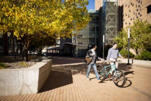 Two people walking with bicycles in a city square, surrounded by trees and modern buildings, engaged in conversation.